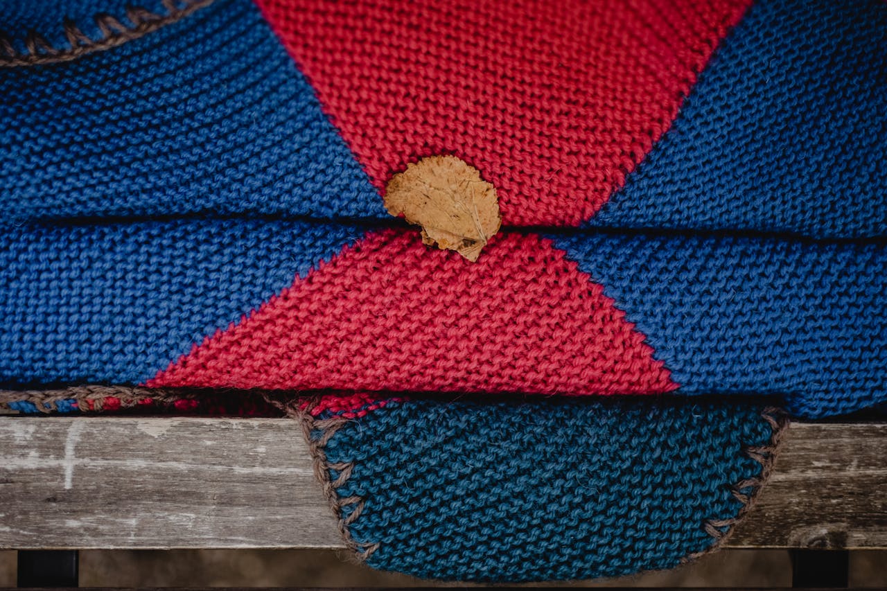 Home Handmade knitted blanket in vibrant red and blue featuring a fallen leaf on a wooden bench.