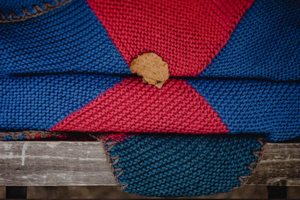 Handmade knitted blanket in vibrant red and blue featuring a fallen leaf on a wooden bench.